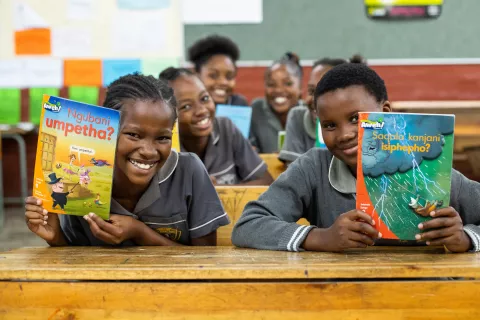 girls smiling broadly, holding up books in their mother tongue that they are reading