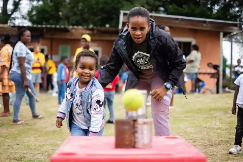 A little girl smiles as she looks at the tennis ball she just threw, aimed at some rusty cans on a table. An older youth is encouraging her from the side.