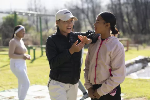 A girl wearing a black jacket and a white cap rests with her elbow on the shoulder of another girl, discussing something.