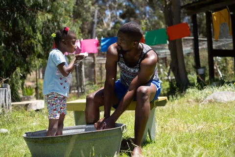 A girl standing in a washing tub blows soap bubbles at her dad who is busy washing clothes in the tub.