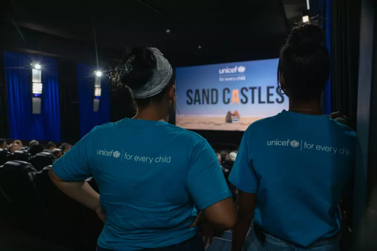 Two UNICEF volunteers wearing UNICEF t-shirts stand at the back of the cinema looking at the beginning of the screening of sand castles.