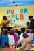 A women wearing a yellow Power of Play t-shirt holds up an open picture book for a group children sitting on the floor in front of her to see.