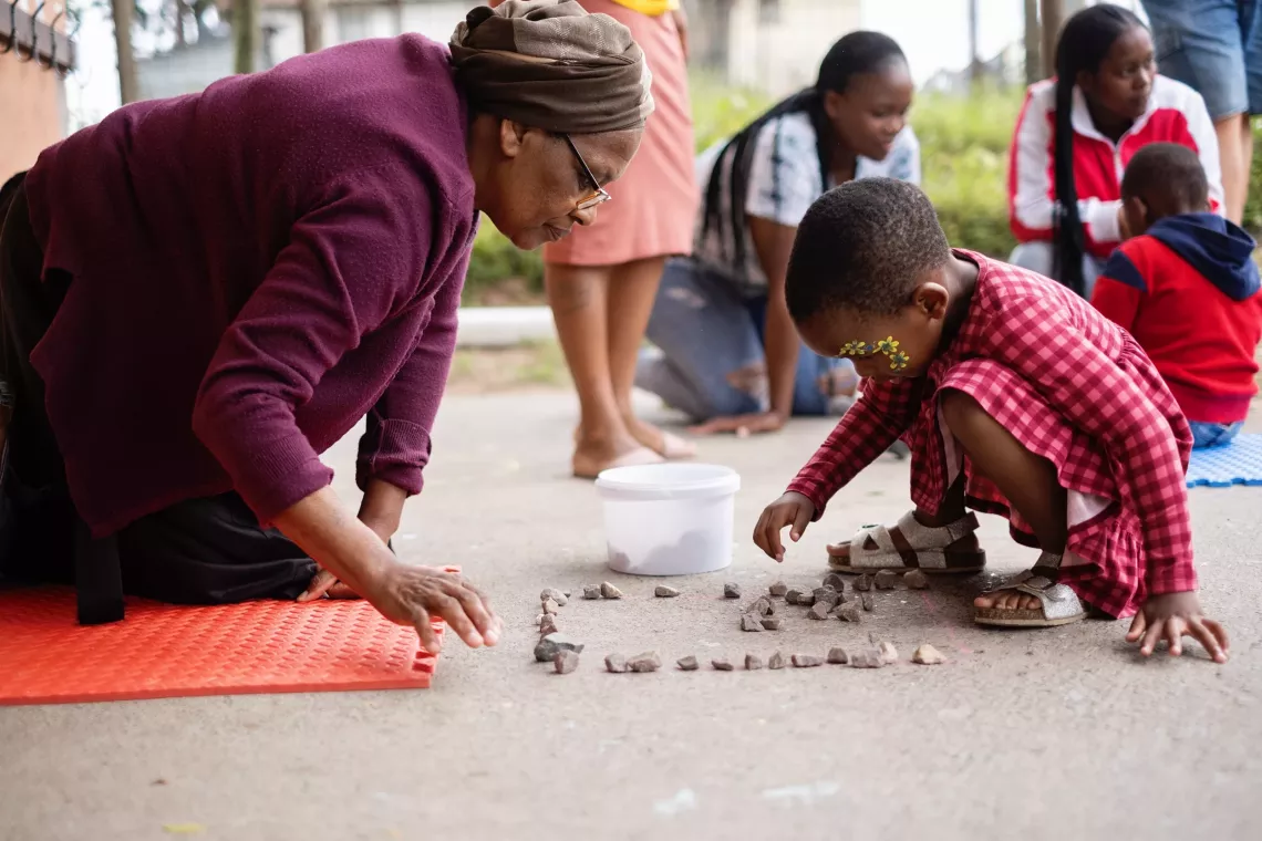 A granny kneels on the ground to look at the patterns that her young granddaughter stacked with small pebbles.