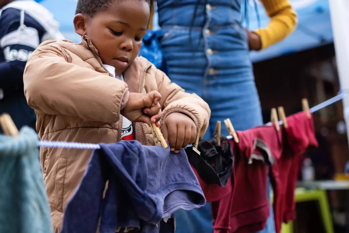 A little boy concentrates on putting a clothes peg to hang a pair of trousers on a low washing line.