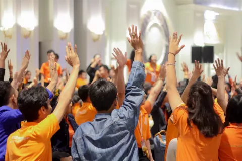 A gathering of young people in a large meeting room, many wearing orange shirts with a few in other colors like blue and black, raising their hands enthusiastically, indicating their participation.