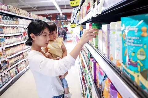 A woman in a white blouse holds a young child in a yellow dress while shopping in a supermarket aisle. She is reaching out to a shelf stocked with various colorful boxes and containers, some marked with sale stickers. The child looks on with a curious expression.