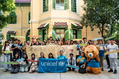 A group of enthusiastic young people gathered outdoors, holding signs with various supportive messages like #Opportunities, #MentalHealth, #Dreams, #Careers, and #Future. They are standing in front of a classic building with large windows and green shutters. At the center is a large banner with the text 'I dream of... for children' and UNICEF banner. To the right, there is a person in a large, friendly UNICEF Teddy Blu mascot. Everyone is smiling, creating a vibrant and hopeful atmosphere.
