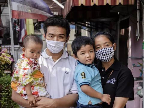 A family photograph. The father is wearing a face mask and is holding his youngest child. The mother, who is also wearing a mask, is holding her child in her arms. Neither of the children is wearing a mask.
