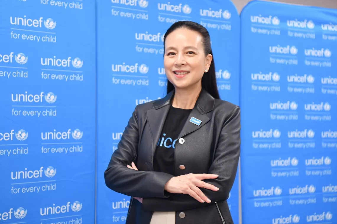 UNICEF Thailand National Ambassadorm, Nualphan Lamsam, stands with her arms crossed in front of a cyan blue graphic backdrop with multiple small, white UNICEF logos.