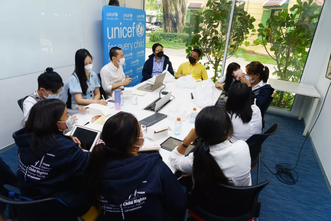 A group of male and female adolescents are gathered in the meeting room of UNICEF. A UNICEF sign is located in the upper left corner of the room.