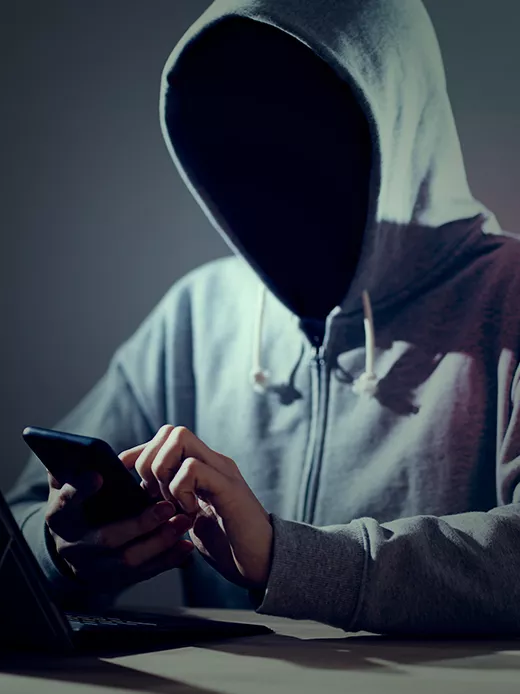 An individual in a dark room, the face hidden by the shadow of a gray hoodie, is engrossed in using a mobile phone. The glow from the screens is the only source of light.