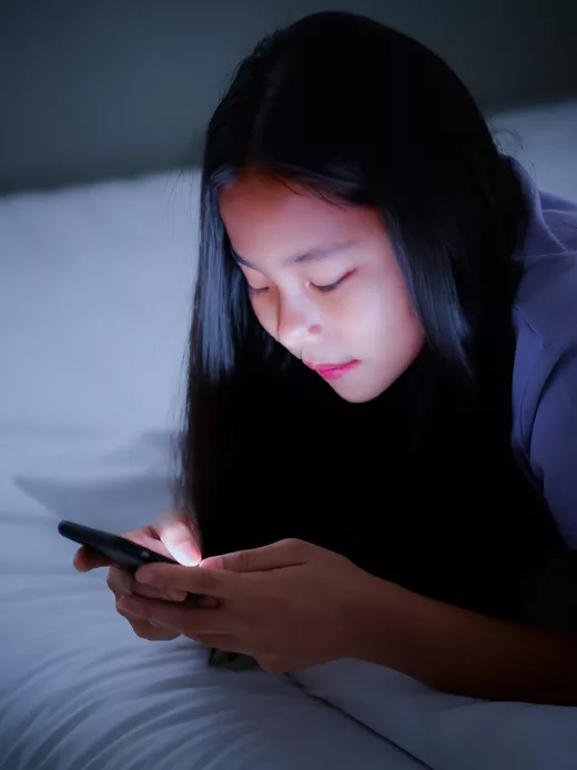 A young girl in a purple shirt is laying on a bed with white sheets, engrossed in texting on a smartphone.
