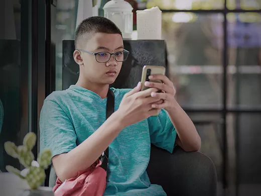 A young boy in a blue t-shirt is sitting on a chair, engrossed in using a smartphone. The background features a window.