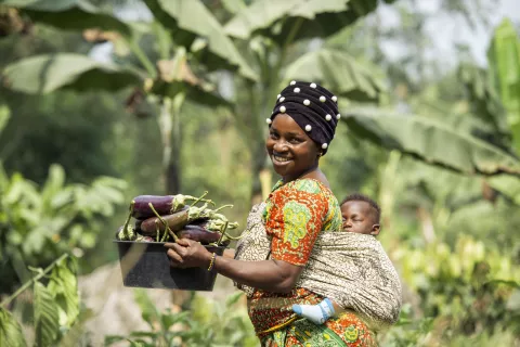 A woman with a basket full of eggplants, carrying her baby sleeping on her back