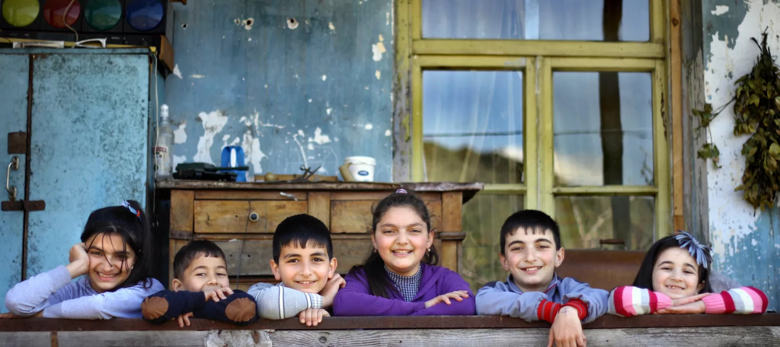 Six children standing behind the fences in front of the old building in the village and putting their arms on the fence and smiling.