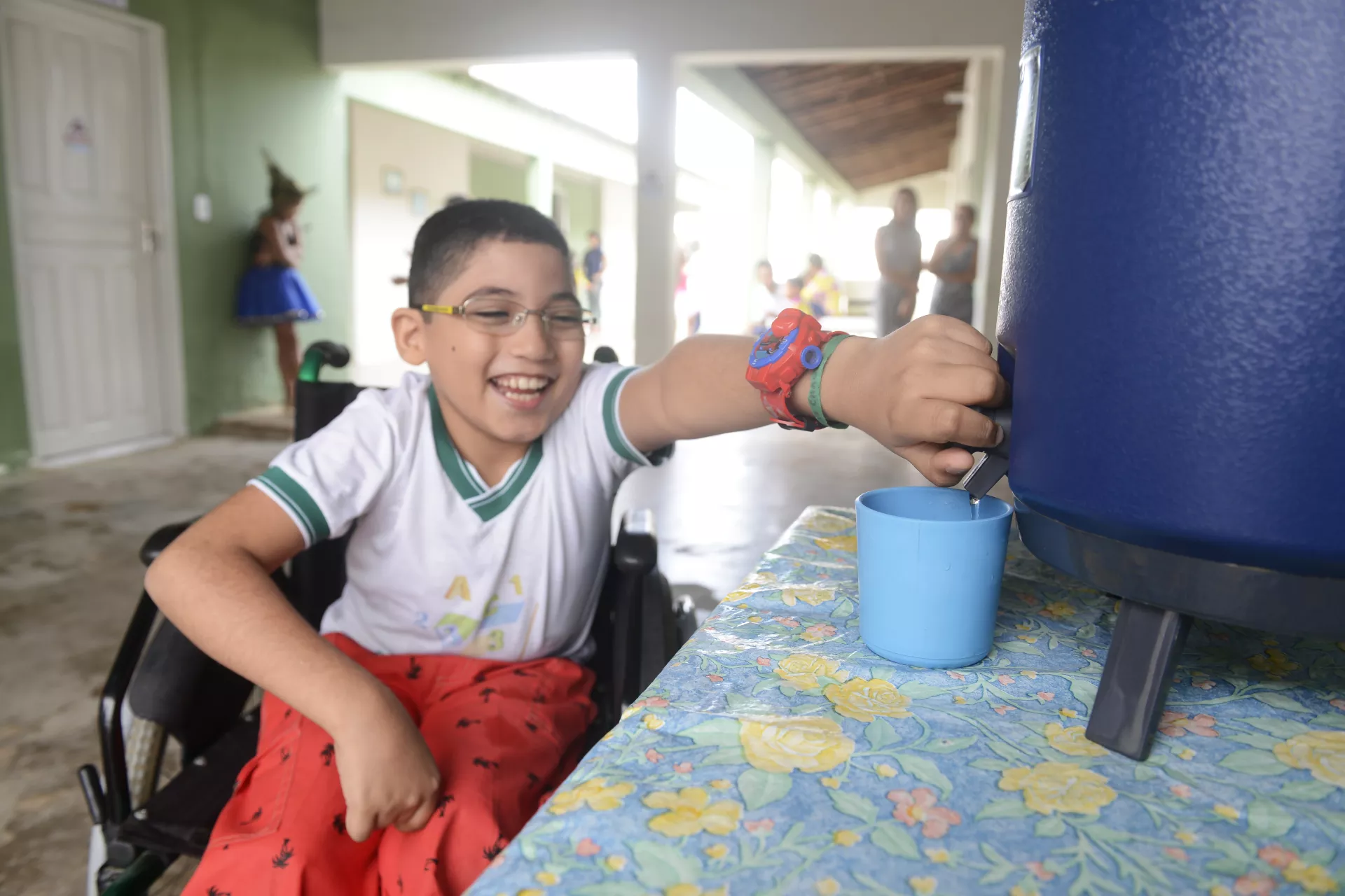 Foto mostra um menino pegando água de um filtro na escola. Ele está sentado em uma cadeira de rodas