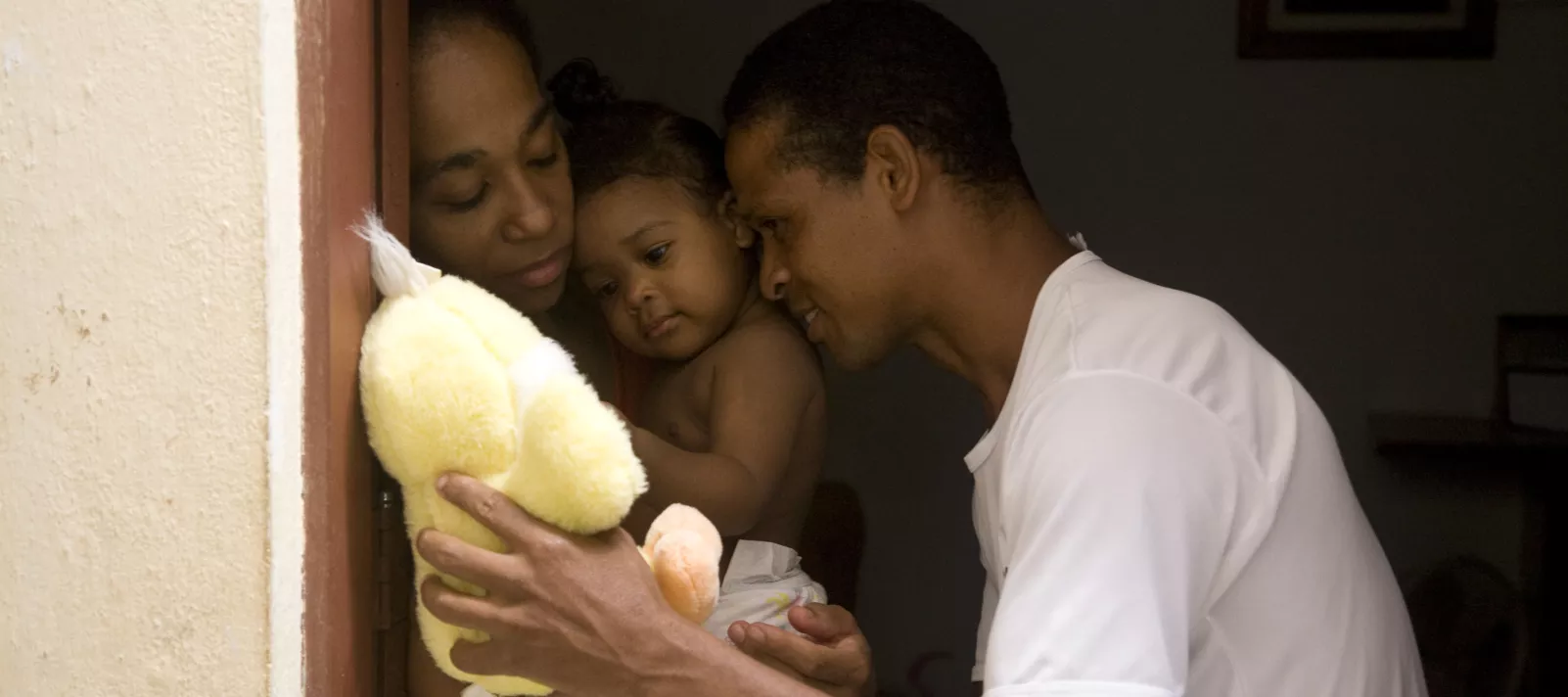 um pai mostra um ursinho de pelúcia amarelo para sua filha bebê que está no colo da mãe. a cena é toda muito carinhosa.