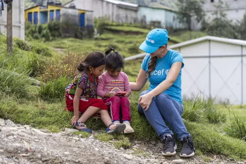 Woman wearing UNICEF branded cap and shirt sitting on the ground and playing with two girls.