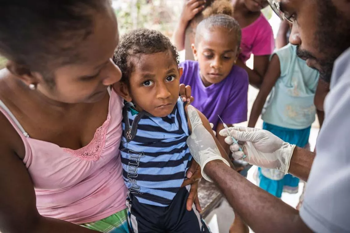 a woman holds a child who is being inoculated by a member of an immunization team that has come by boat from Port Vila, the capital, to vaccinate children on Ifira Island