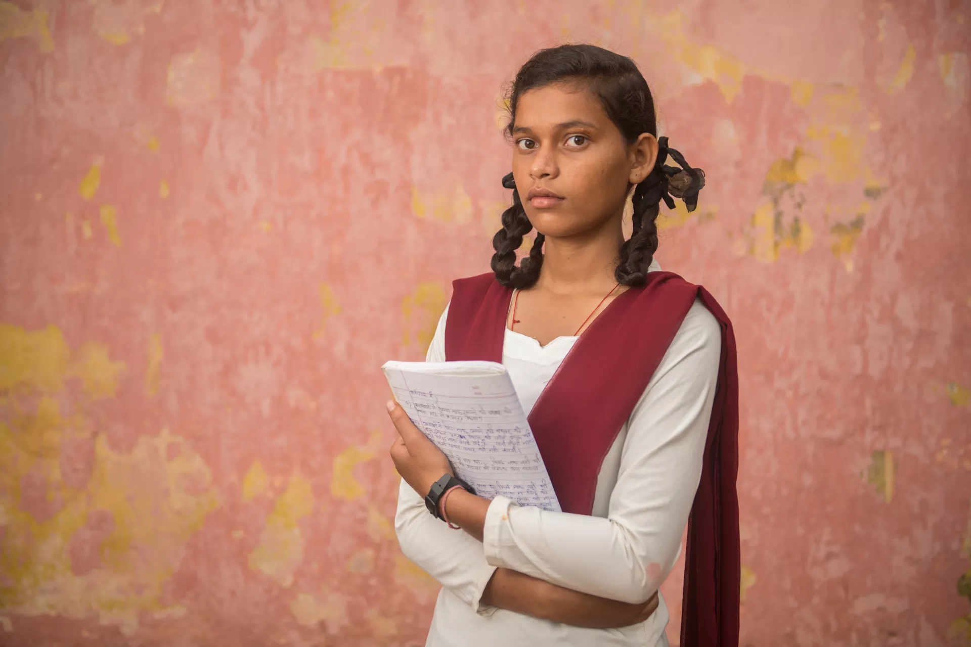 An adolescent girl, Kusma, looks into the camera with determination while holding a school book against a salmon-colored wall.
