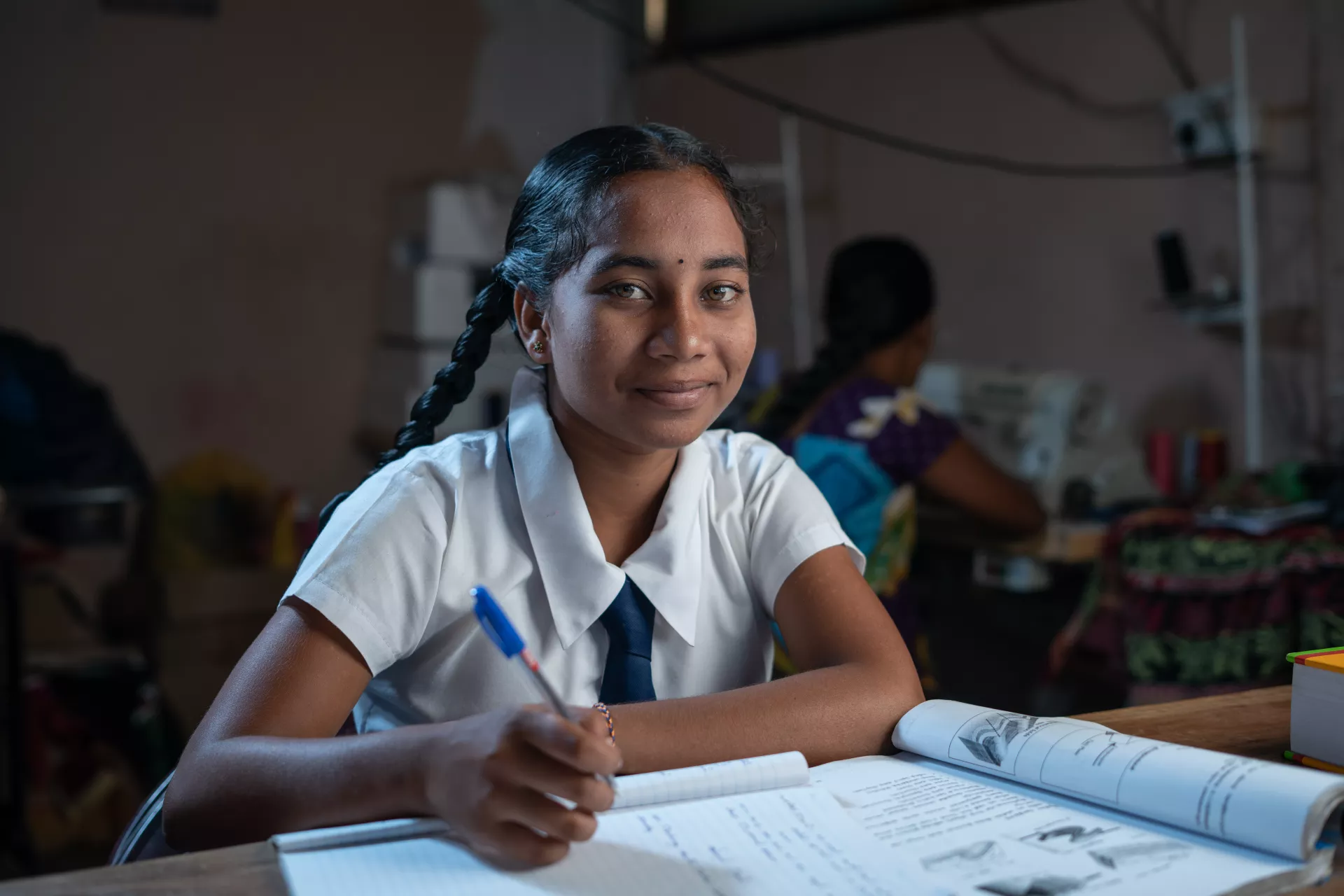 Yathusia looks confidently at the camera in her school uniform: braids (black hair), a white button-up and black tie. On the desk in front of her a textbook and notebook are open and pen is in her hand. In the dark, blurred background another student is sitting with her back to the camera.