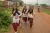 : Kusma runs down an dirt road with three other school girls, though one is barely visible behind another. They wear off white kurthis and maroon pants. Their hair is tied in braids with pink ribbons. Behind them, on either side of the road are scenes of the village and a motorbike drives down the road. 