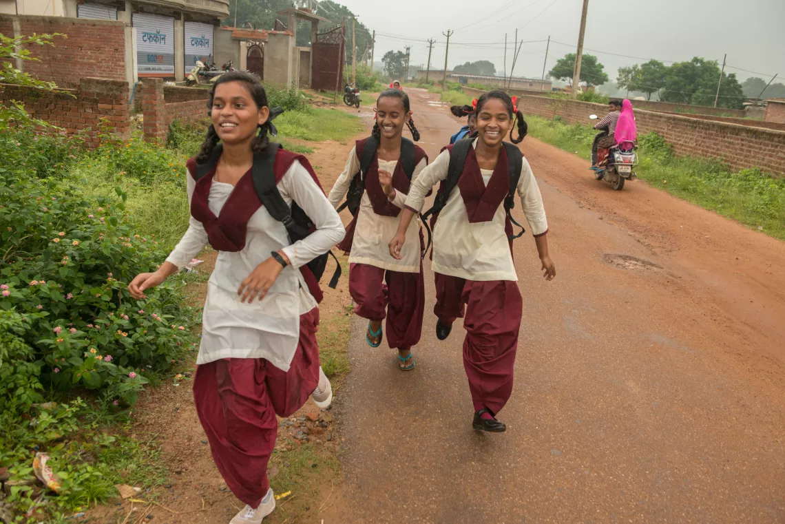 : Kusma runs down an dirt road with three other school girls, though one is barely visible behind another. They wear off white kurthis and maroon pants. Their hair is tied in braids with pink ribbons. Behind them, on either side of the road are scenes of the village and a motorbike drives down the road. 