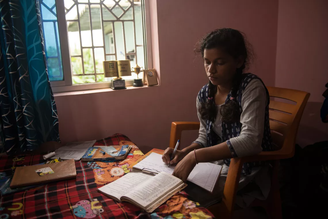 Kusma is studying indoors next to a window, with daylight streaming in. She writes in a notebook, in her lap, while looking at a textbook on top of a table. 
