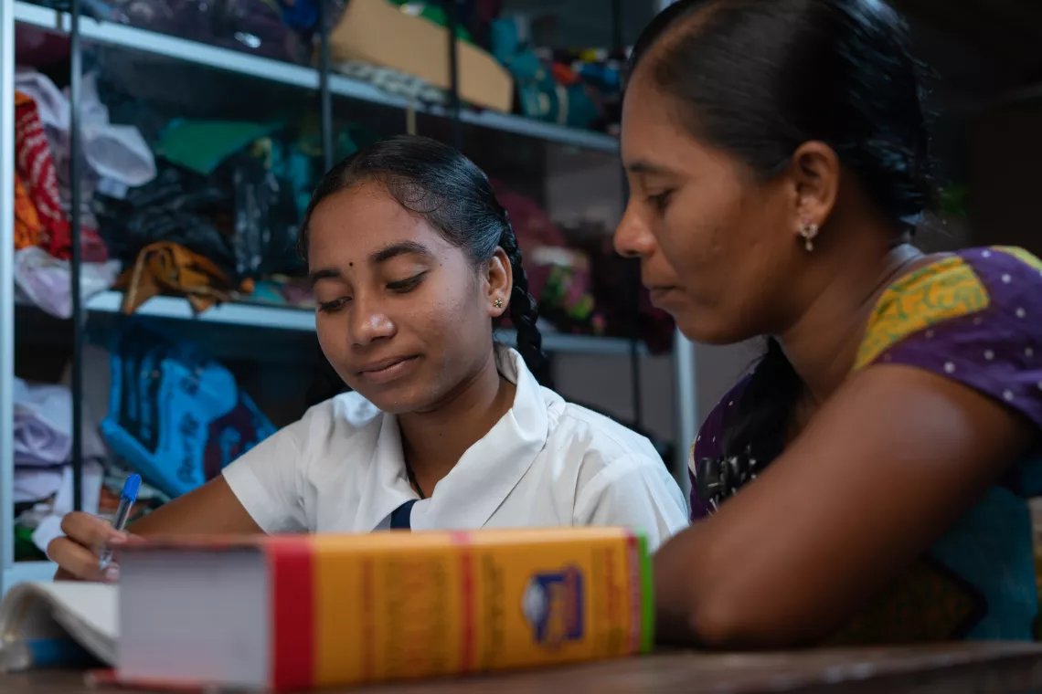 Yathusia studies with her mom. They read from a textbook together. In the blurred background are shelves with clothes.