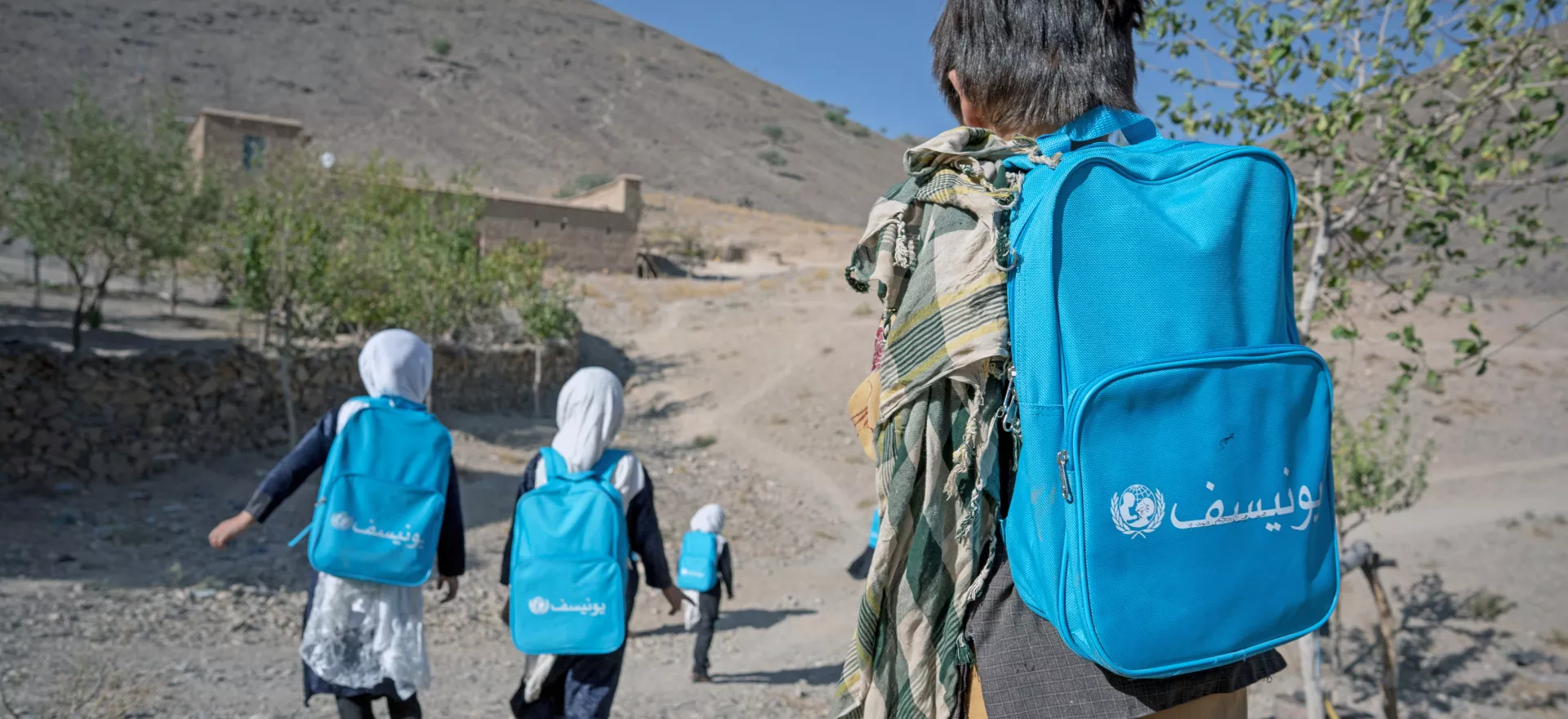 Vue de dos. Des enfants portant un sac à dos UNICEF marchent sur une route de montagnes.