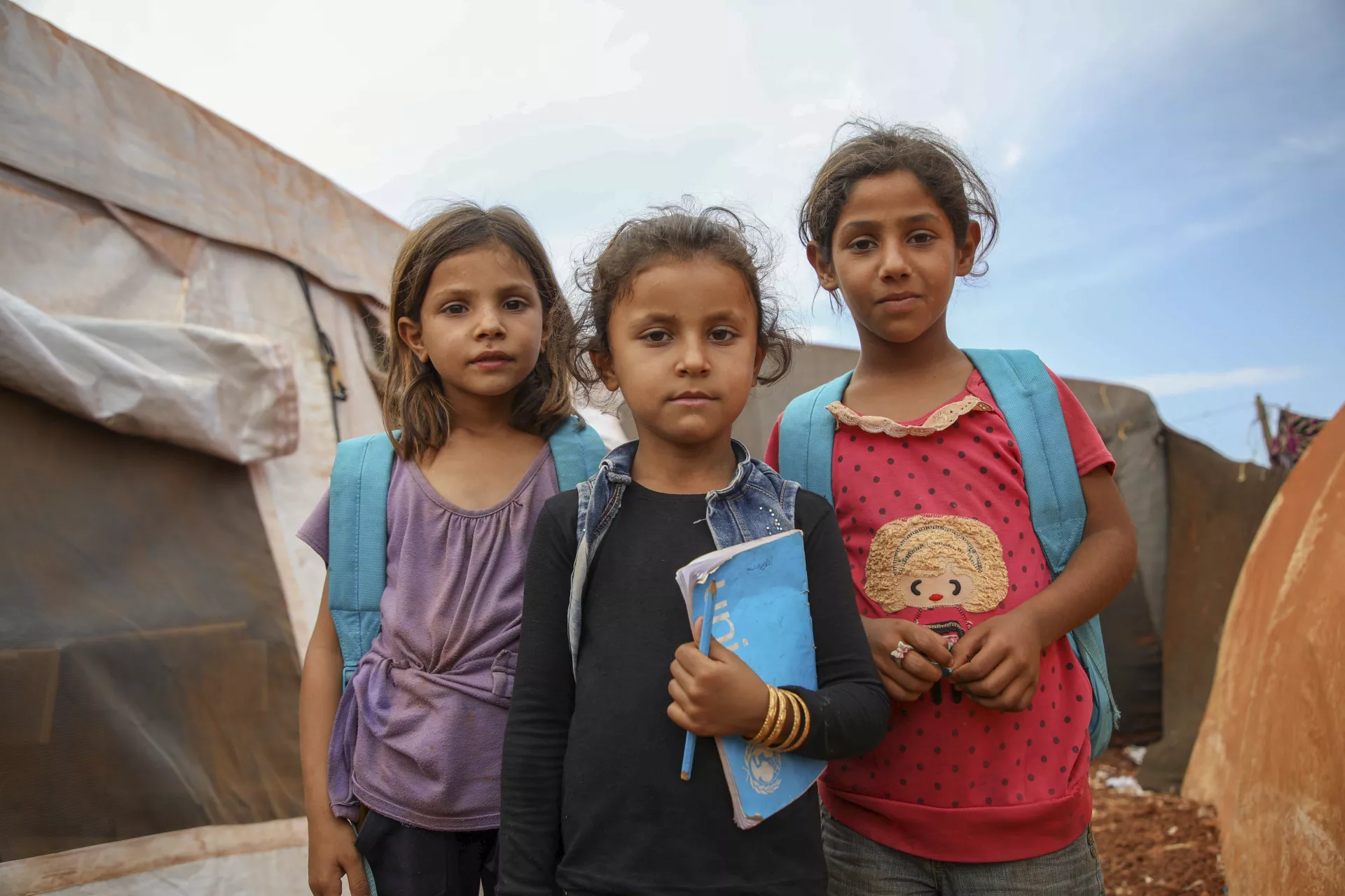Three girls in backpacks stand outside a tent school in a makeshift camp in the Syrian Arab Republic.