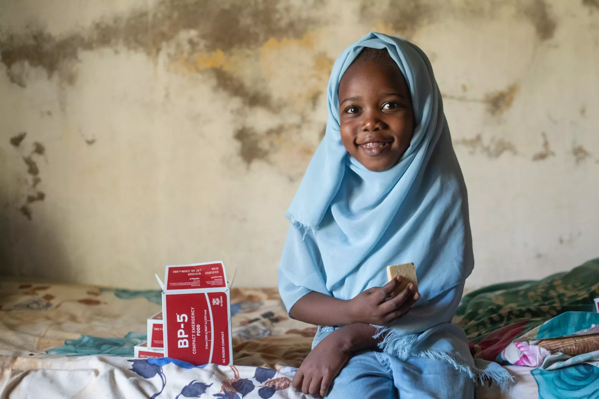 A young girl in a blue scarf nibbles on fortified biscuits provided during an integrated health campaign in Gezira State, Sudan.