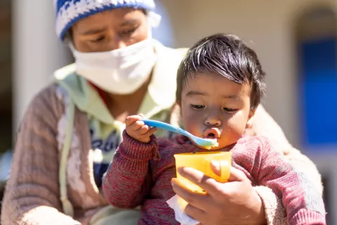 Guatemala. A small child eats mashed fruit from a bowl while being held by his mother.