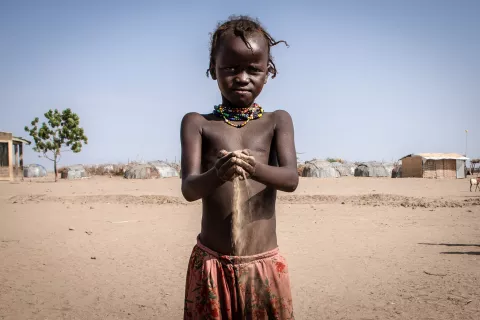 Un enfant se tient debout, laissant couler du sable entre ses doigts. Il se trouve dans un paysage aride