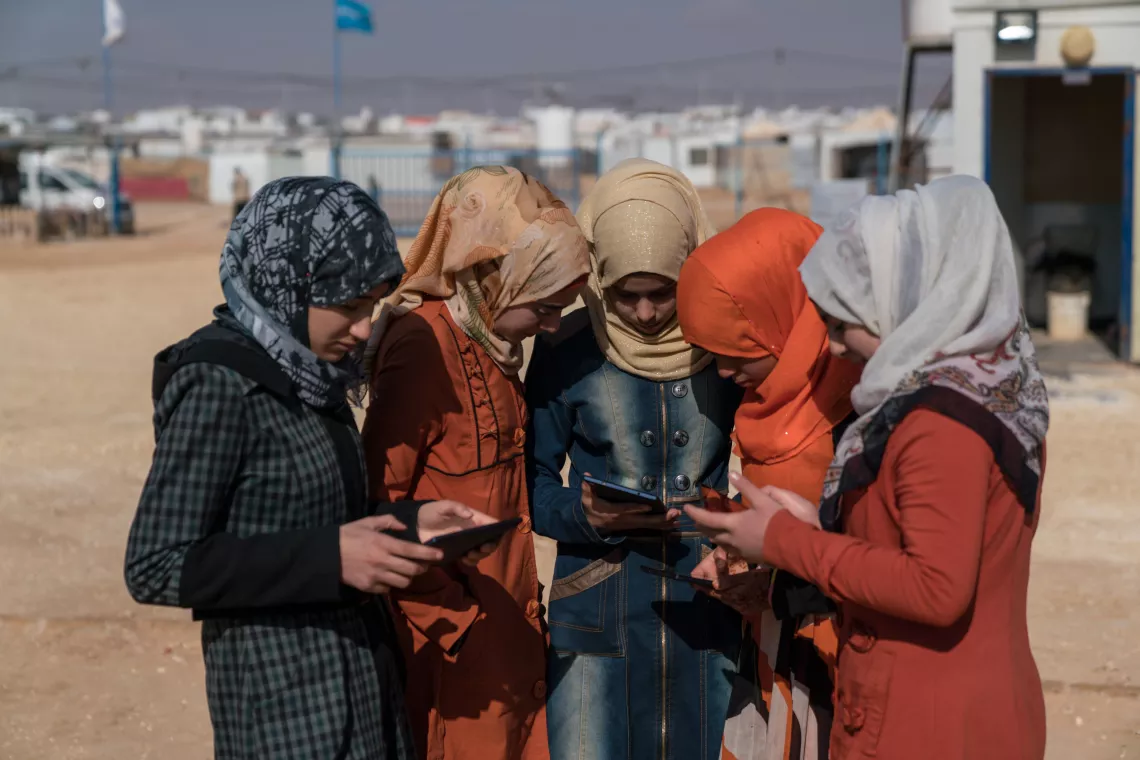 Five girls stand outside in a refugee camp, looking at cell phones and tablets.