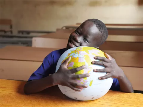 A boy sitting at a school desk holds a plastic globe. 