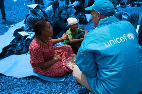 A male staff member wearing a jacket bearing the UNICEF logo talks with a woman sitting on the ground. Close to her, several other people sitting on the ground watch the conversation. 