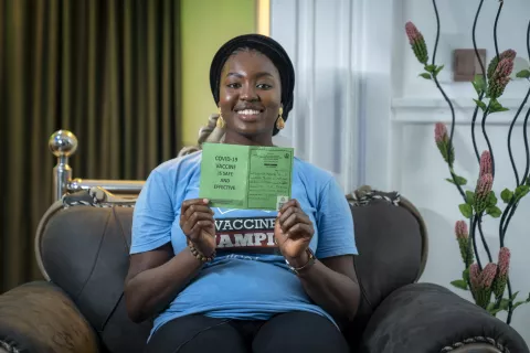 Young woman showing her COVID-19 vaccine card to the camera, while smiling and sitting on a couch